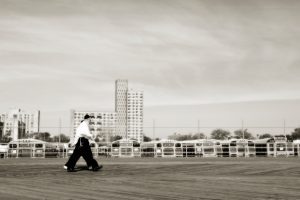 Photograph of two men walking across the boardwalk on Coney Island in front of a school bus station.