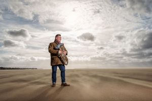 Musician Gary Curtin photographed on Hoylake beach holding his euphonium. Wind is blowing sand across the flat beach and the sun is shining through the clouds behind him.
