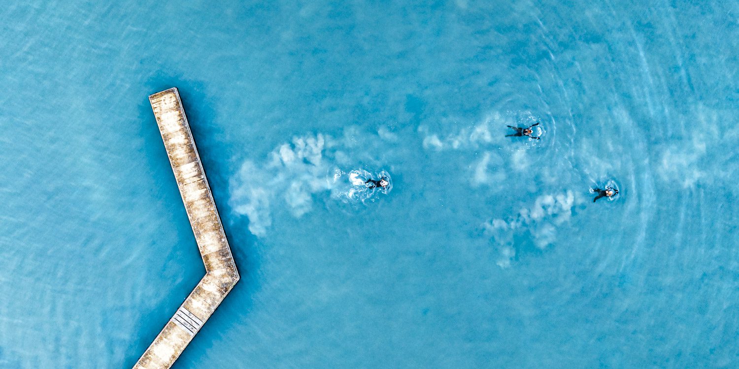 Top down drone image showing a jetty to the left of the picture and then three swimmers in wetsuits swimming away from the jetty to the right of the picture.