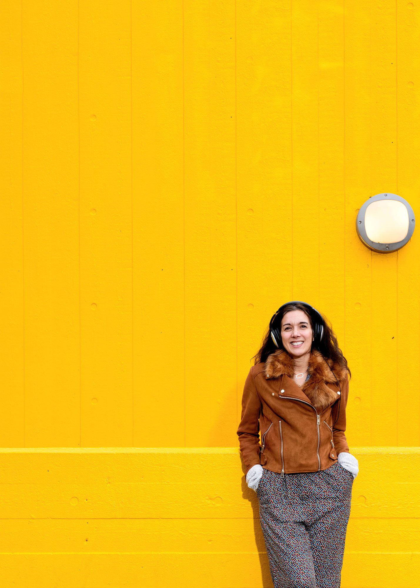 Woman wearing headphones leaning against a bright yellow walls.