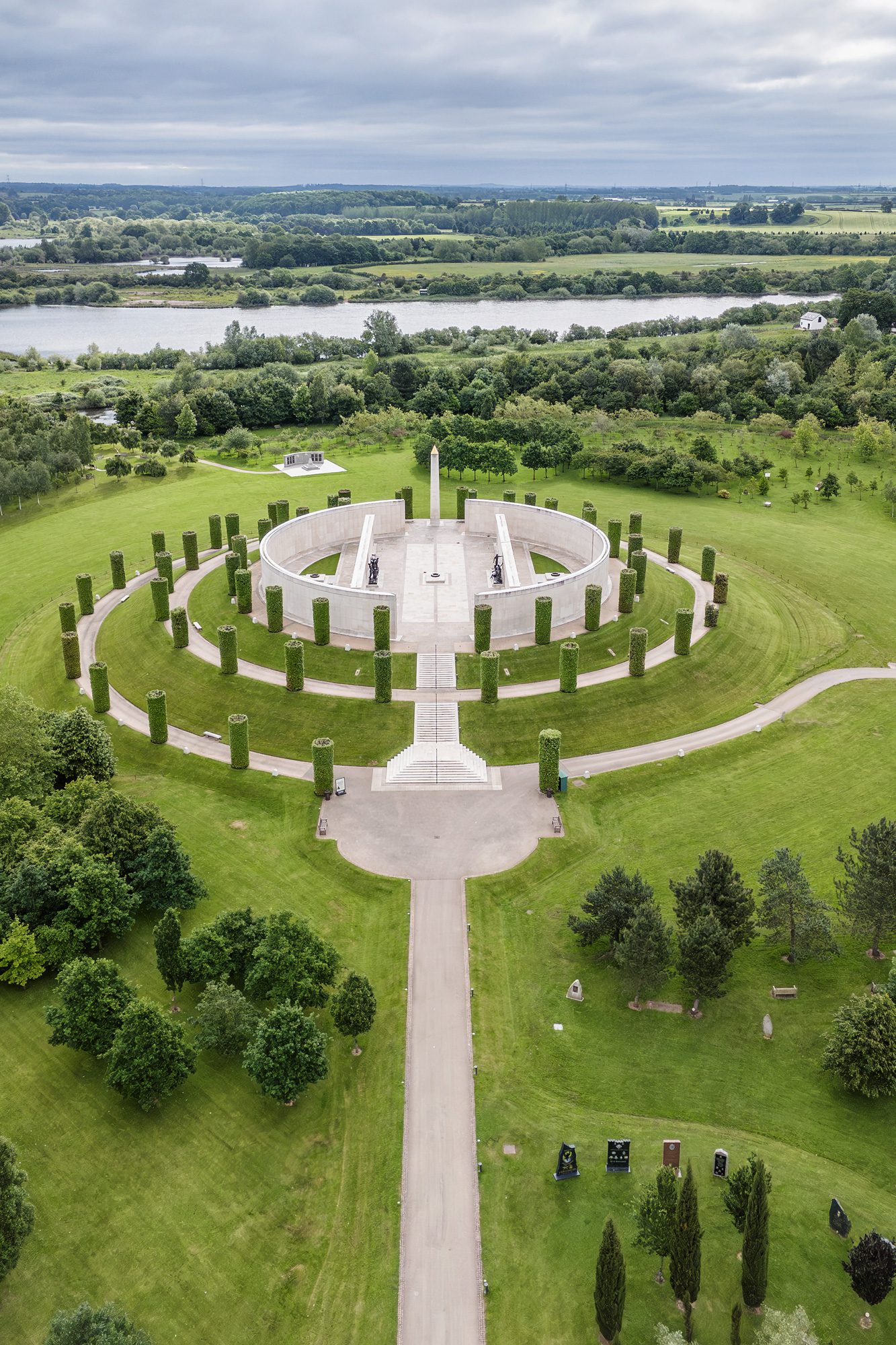 Aerial photograph of the Armed forces memorial which is within a stone circle on the top of a small hill surrounded by a spiralling path lined with trees. Byrne the memorial is a river and then countryside.